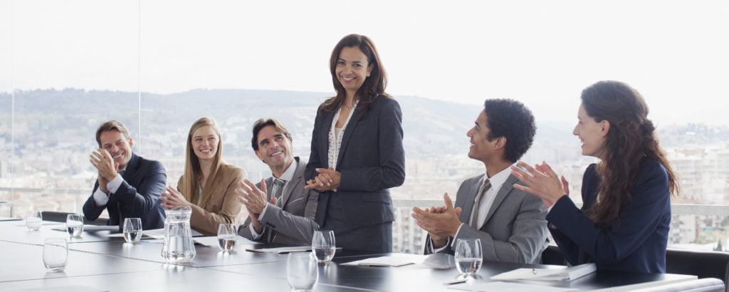 woman standing at conference room table and team members clapping