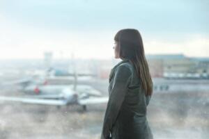 A woman in a gray jacket looks out an airport window at the runway and parked airplanes. The scene is blurred and overcast, creating a contemplative mood.